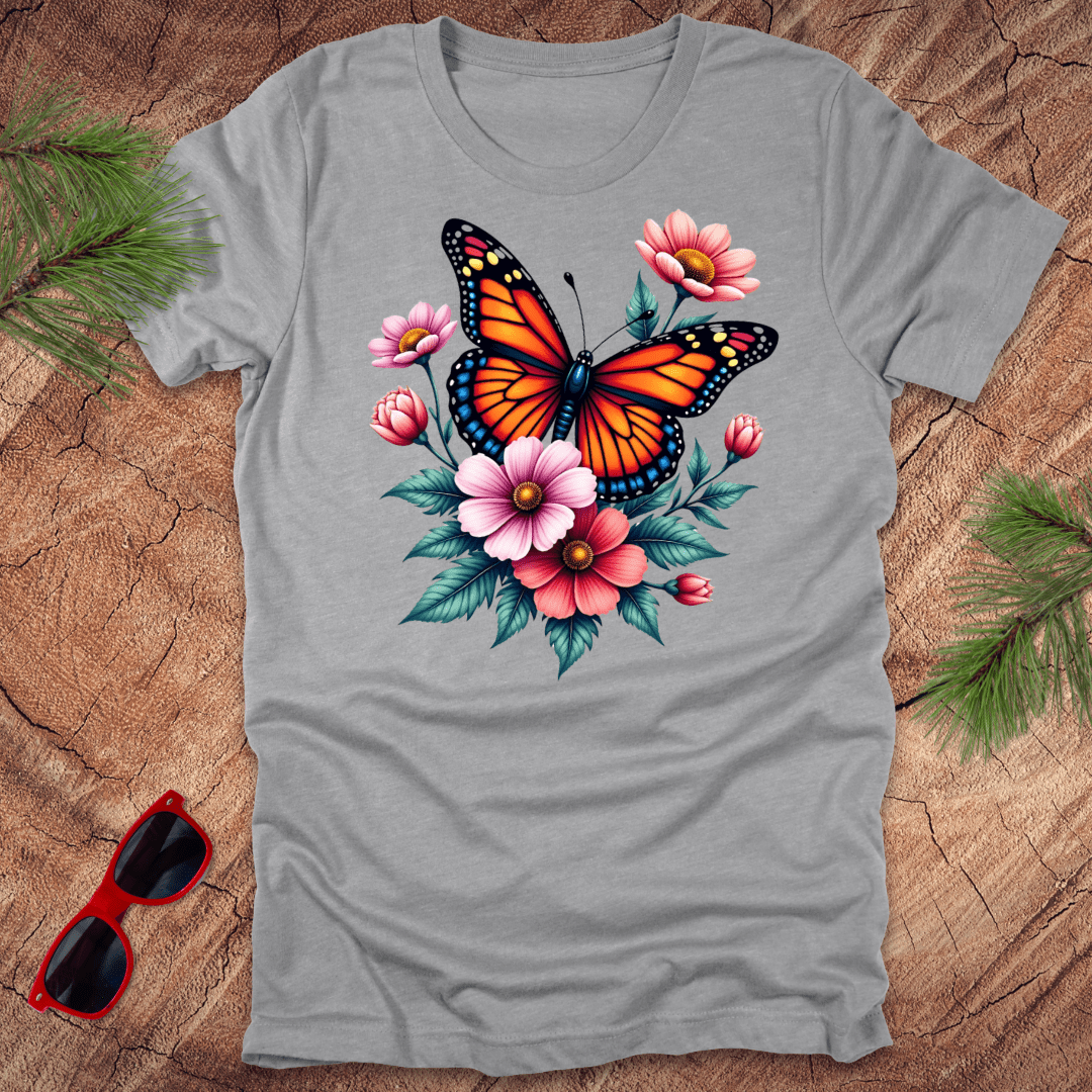 A gray t-shirt with a vibrant print of a butterfly surrounded by pink and red flowers, displayed on a wooden surface with green leaves and red sunglasses to the side.