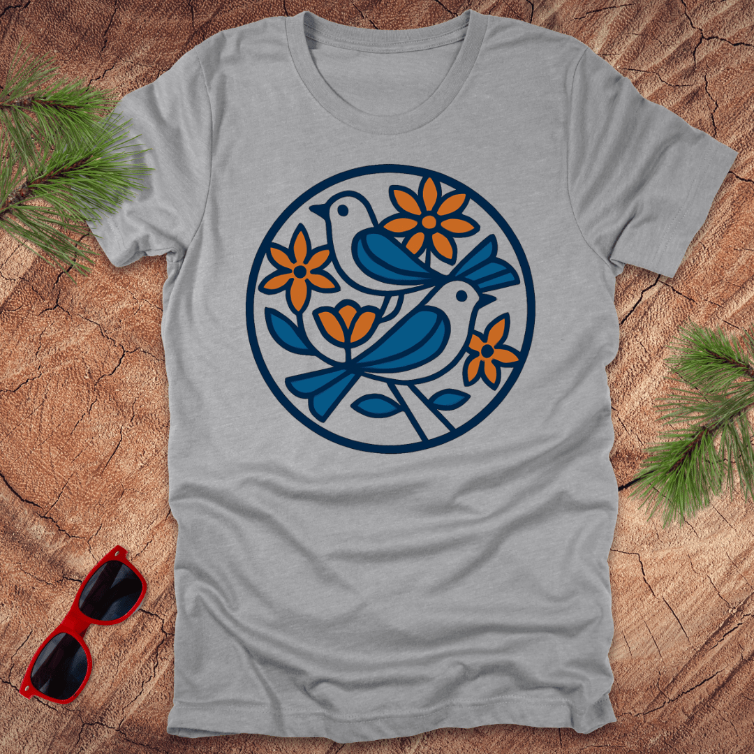 Gray t-shirt with a blue and orange bird and flower design on a wooden surface with sunglasses and greenery.