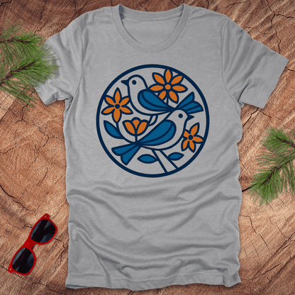 Gray t-shirt with a blue and orange bird and flower design on a wooden surface with sunglasses and greenery.