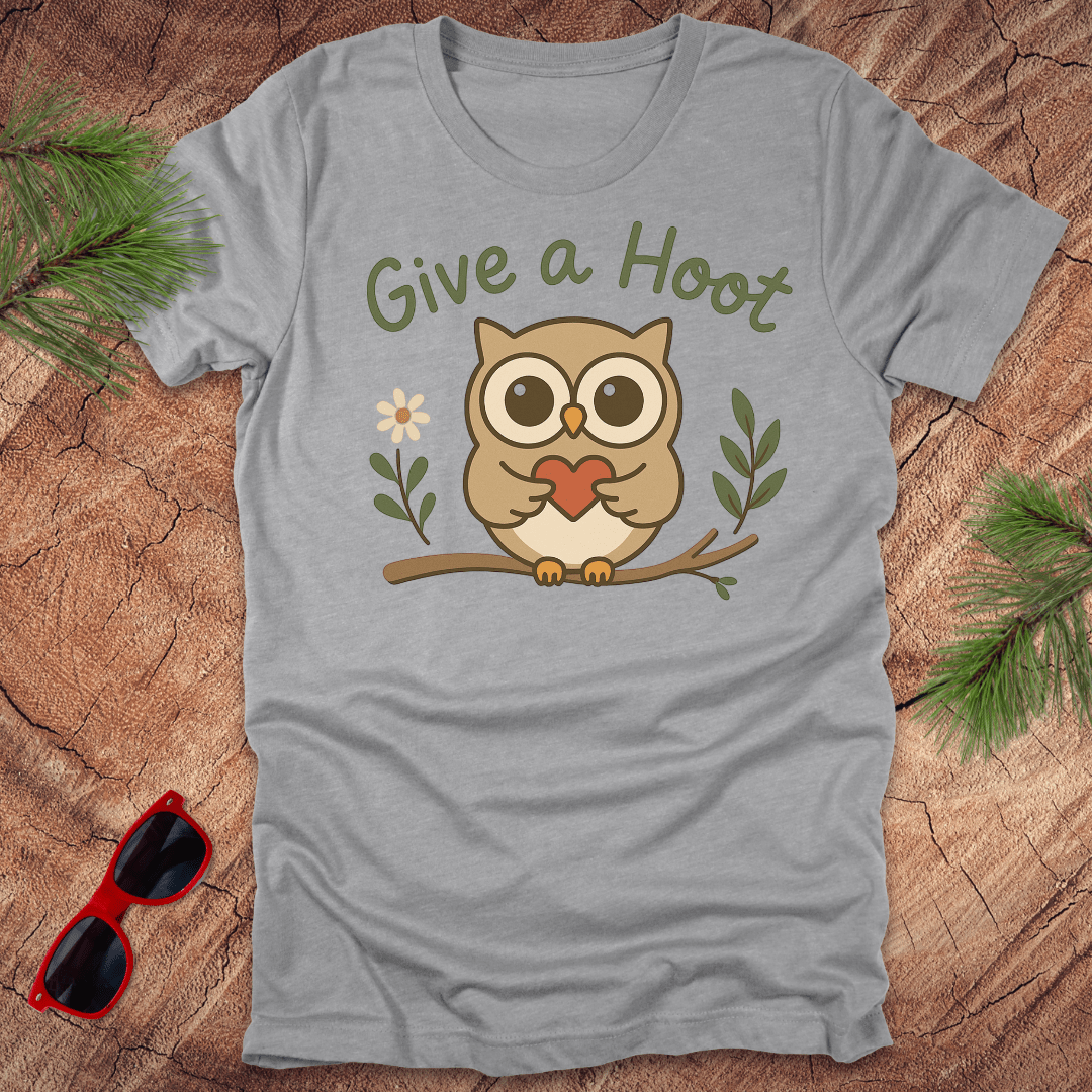 Gray t-shirt with an owl graphic and 'Give a Hoot' text on a wooden surface with sunglasses and greenery.