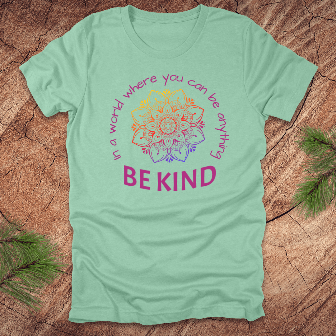 Light green t-shirt with a colorful mandala and 'Be Kind' text on a wooden surface.