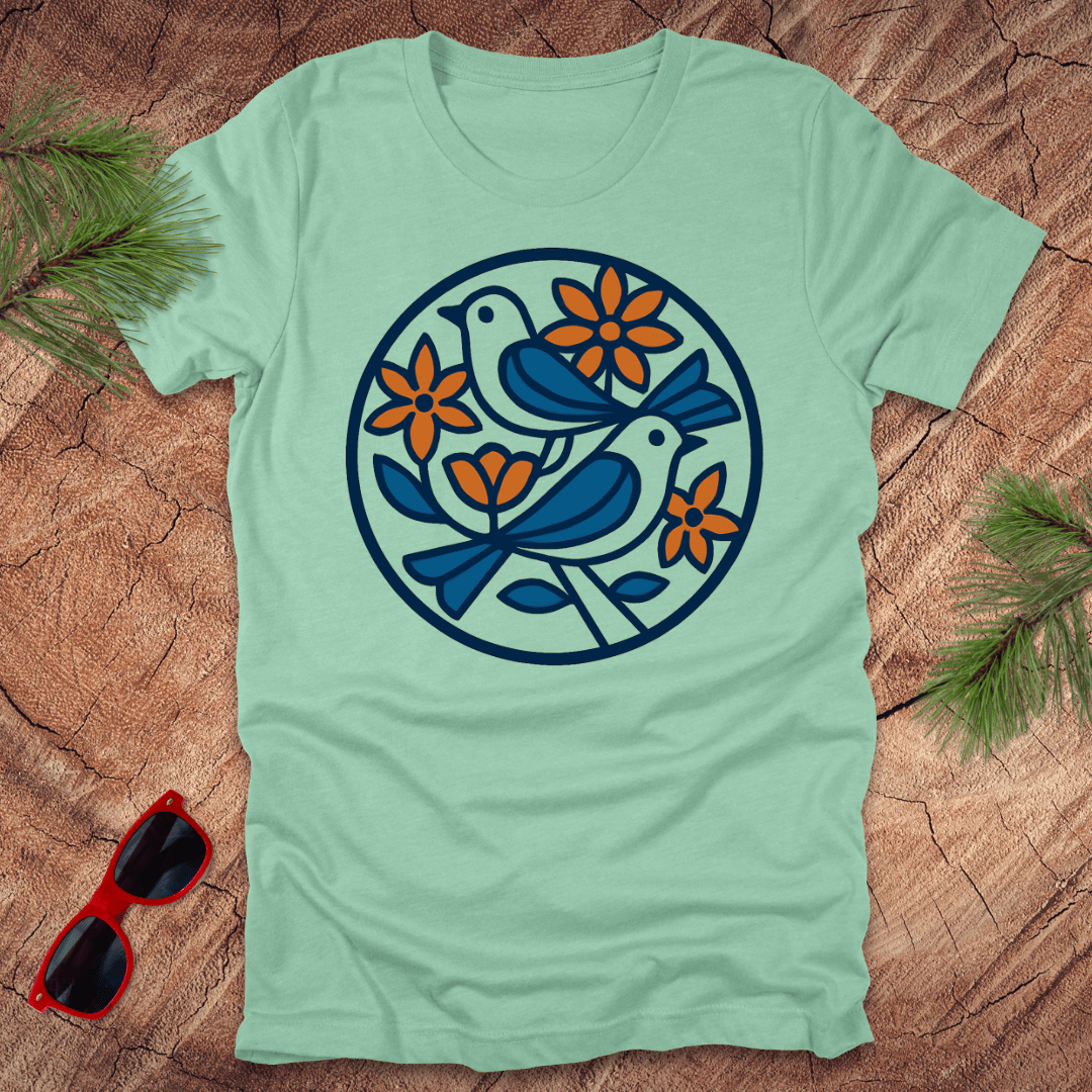Light green t-shirt with bird and flower design on a wooden surface with sunglasses and greenery.