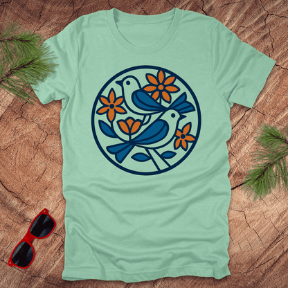 Light green t-shirt with bird and flower design on a wooden surface with sunglasses and greenery.