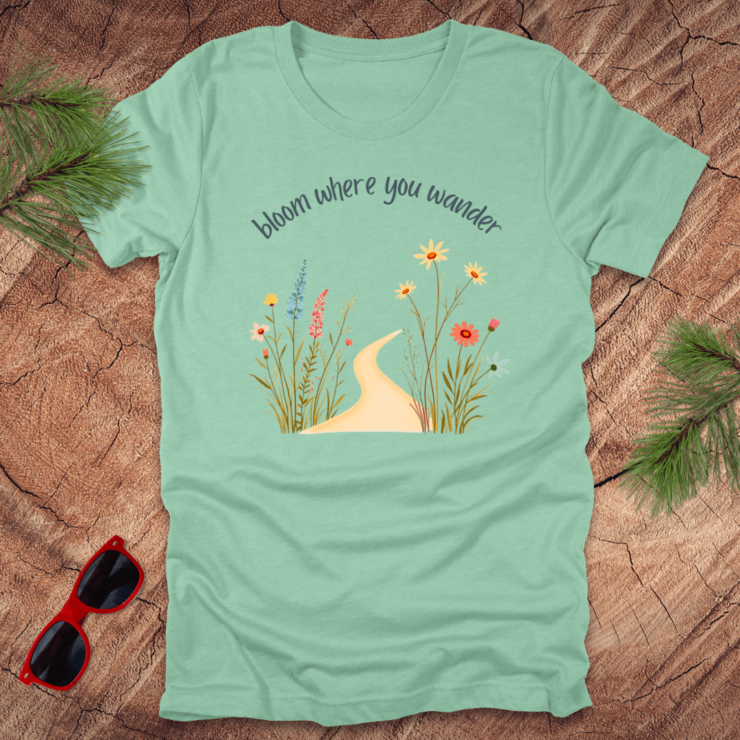 mint green t-shirt with a graphic that includes a floral design and the text 'bloom where you wander', accompanied by  flowers on a wooden background with pine needles and red sunglasses to the side.