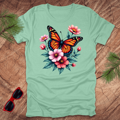 A mint green t-shirt with a vibrant print of a butterfly surrounded by pink and red flowers, displayed on a wooden surface with green leaves and red sunglasses to the side.