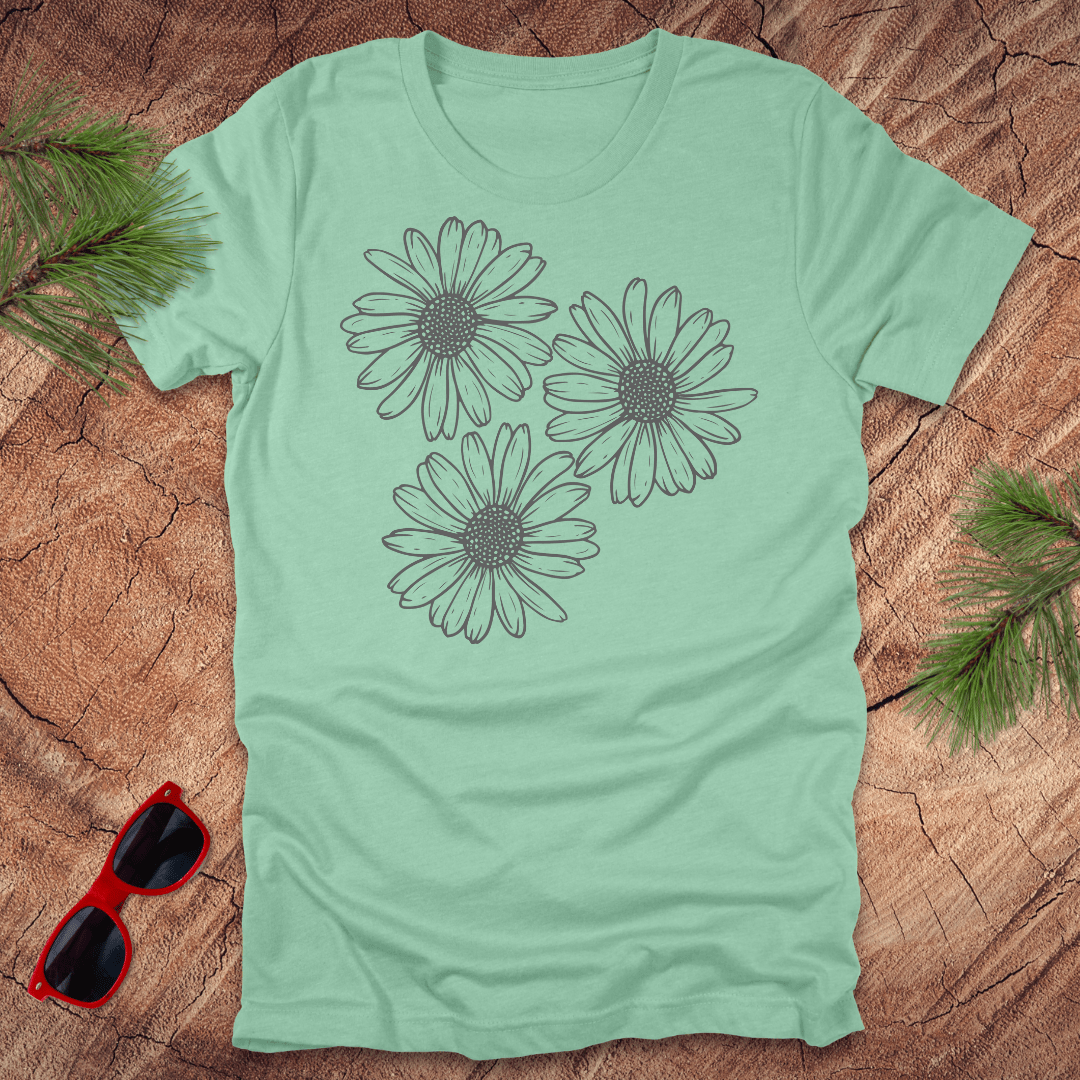 A mint green t-shirt with 3 daisies prominently displayed. on a wooden surface with sunglasses and greenery.