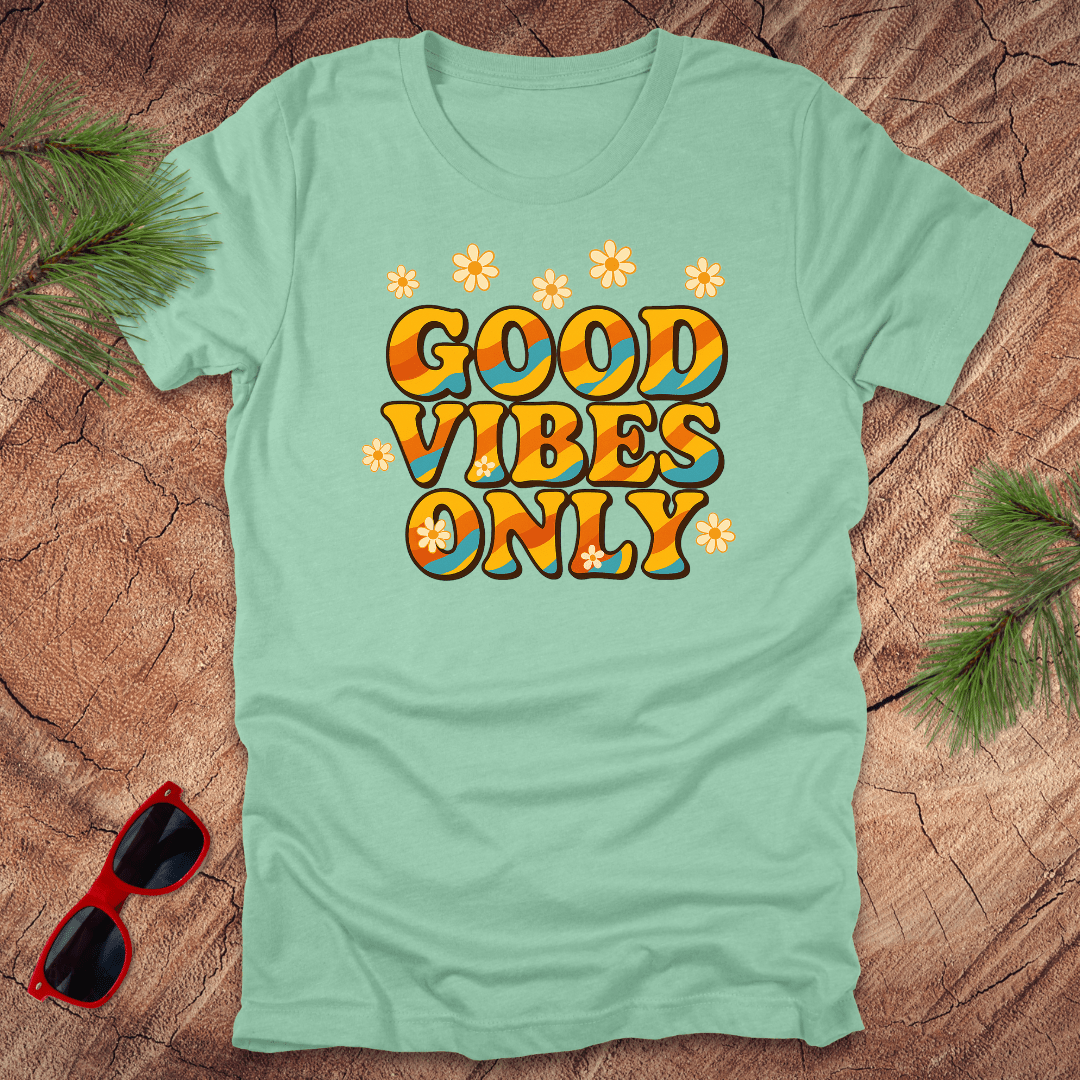 Light green t-shirt with 'Good Vibes Only' text on a wooden surface with sunglasses and leaves.