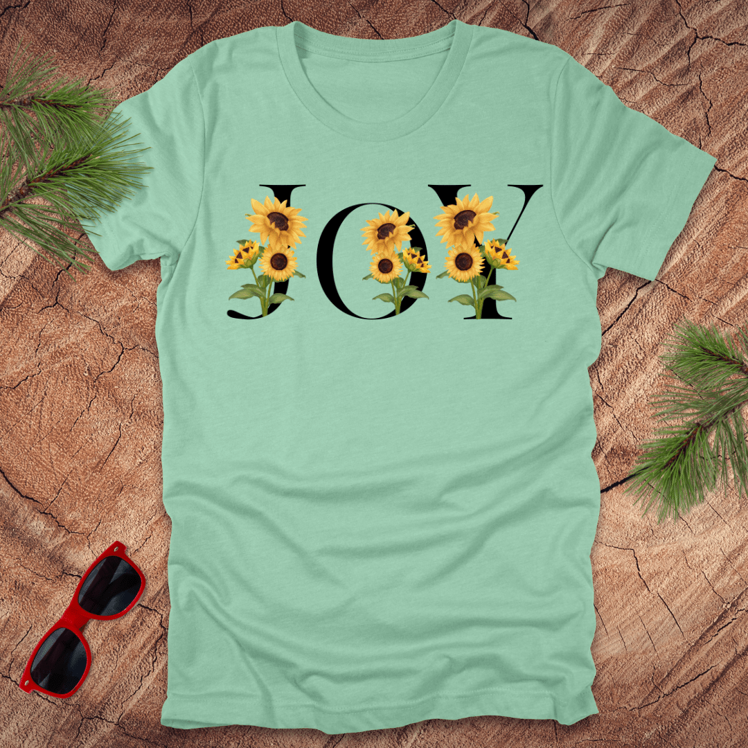 Mint green t-shirt with 'JOY' and sunflower design on a wooden surface with sunglasses and greenery.