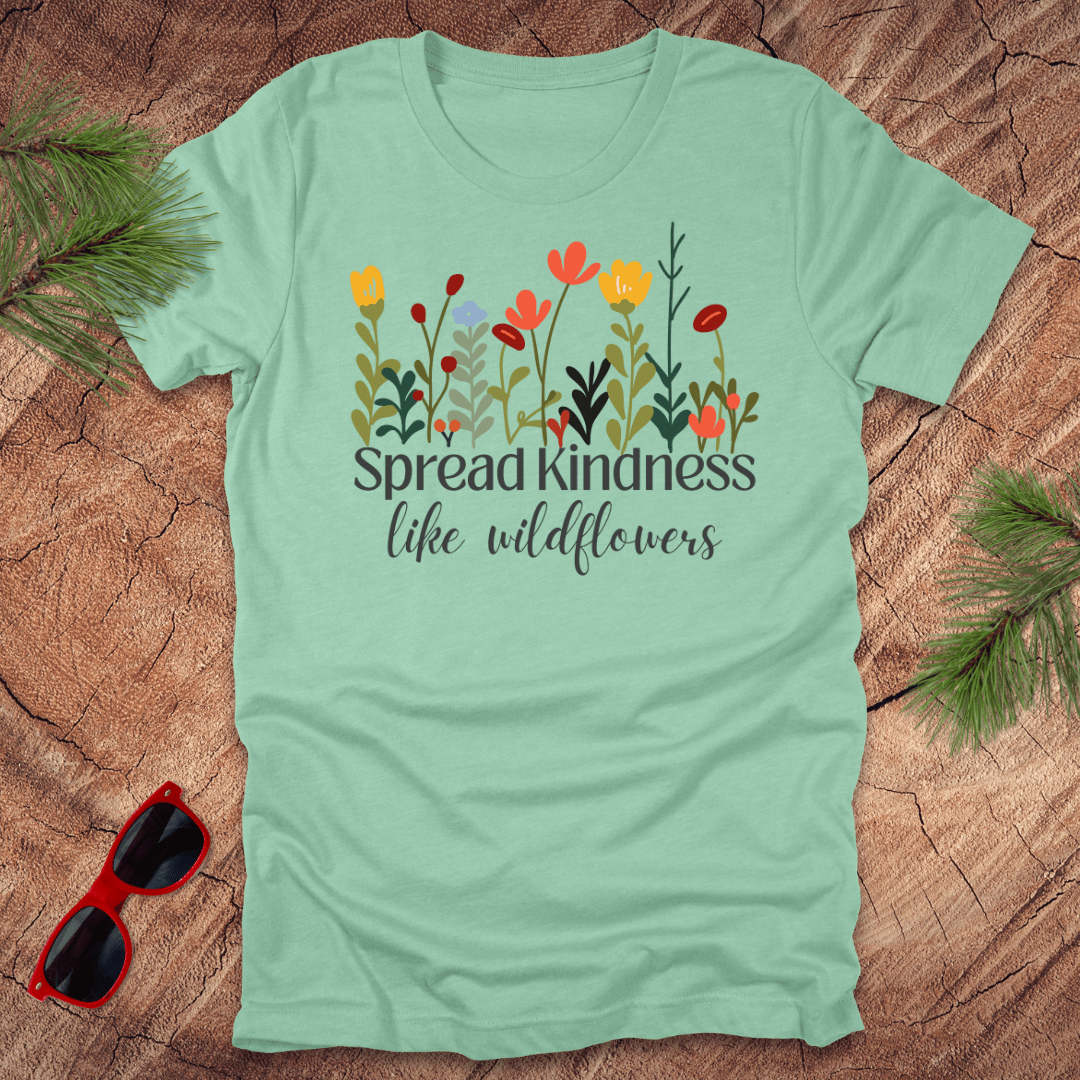 Mint green t-shirt with floral design and text 'spread kindness like wildflowers' on a wooden surface with sunglasses and greenery.