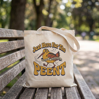 Tote bag with a woodcock graphic and text 'Just Here For The Peent' on a wooden bench outdoors.
