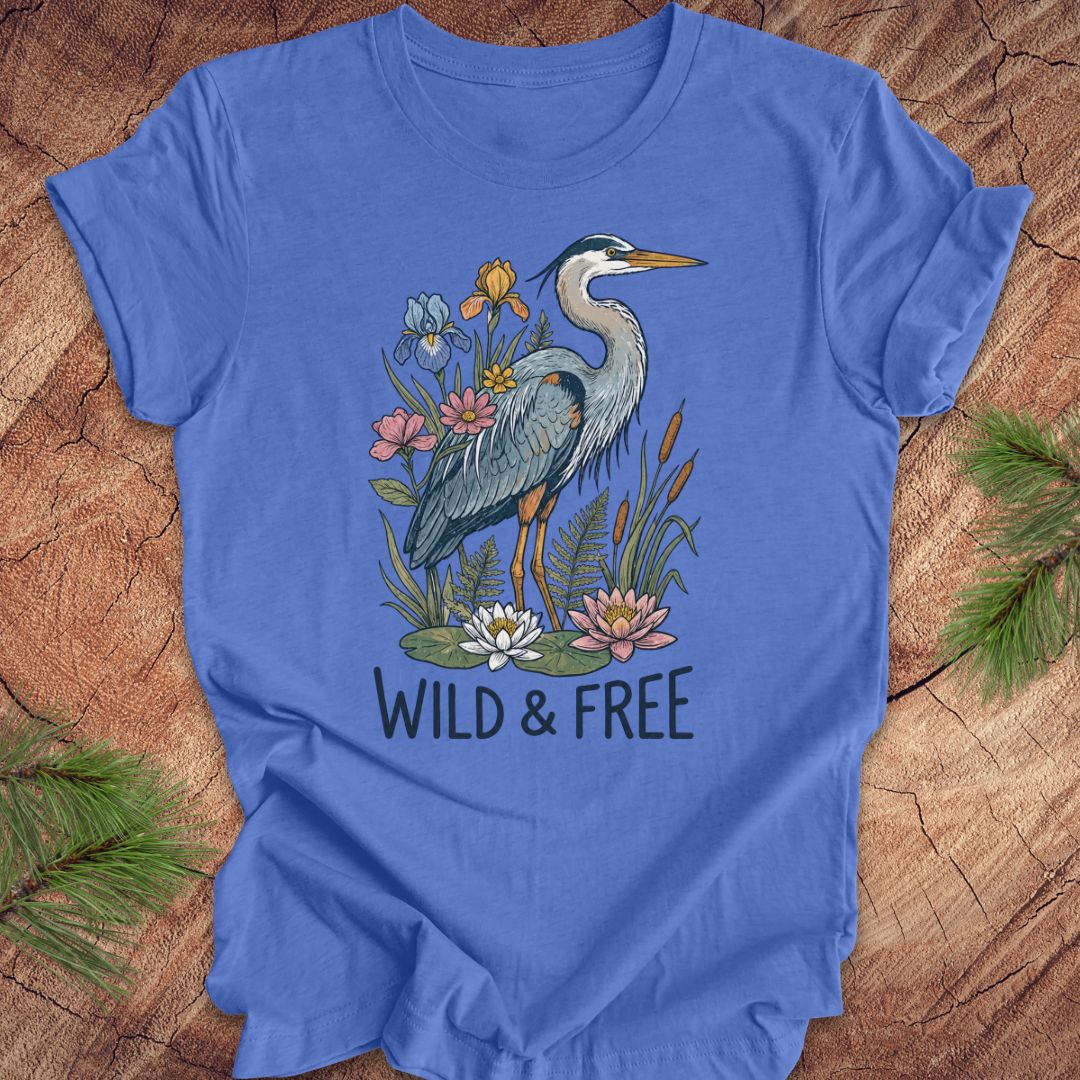 A blue short-sleeve t-shirt with a graphic of a heron surrounded by flowers and the text 'WILD & FREE', laid flat on a rustic wood background with pine branches in the corners.