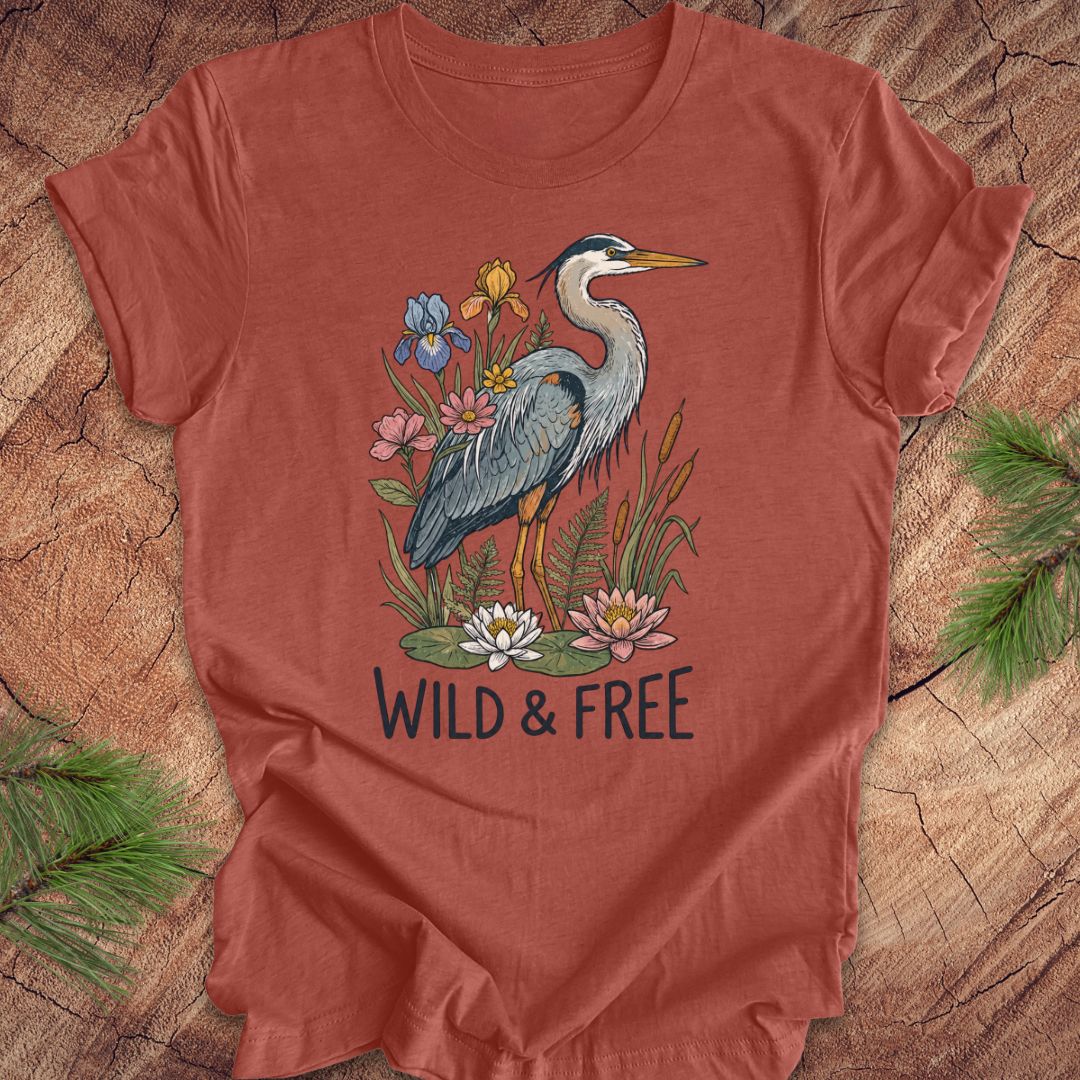 A terracotta-colored short-sleeve t-shirt featuring a detailed graphic of a blue-gray heron surrounded by colorful wildflowers and the text 'WILD & FREE' printed below. The t-shirt is laid flat on a rustic wood slice background with pine needles in the corners.