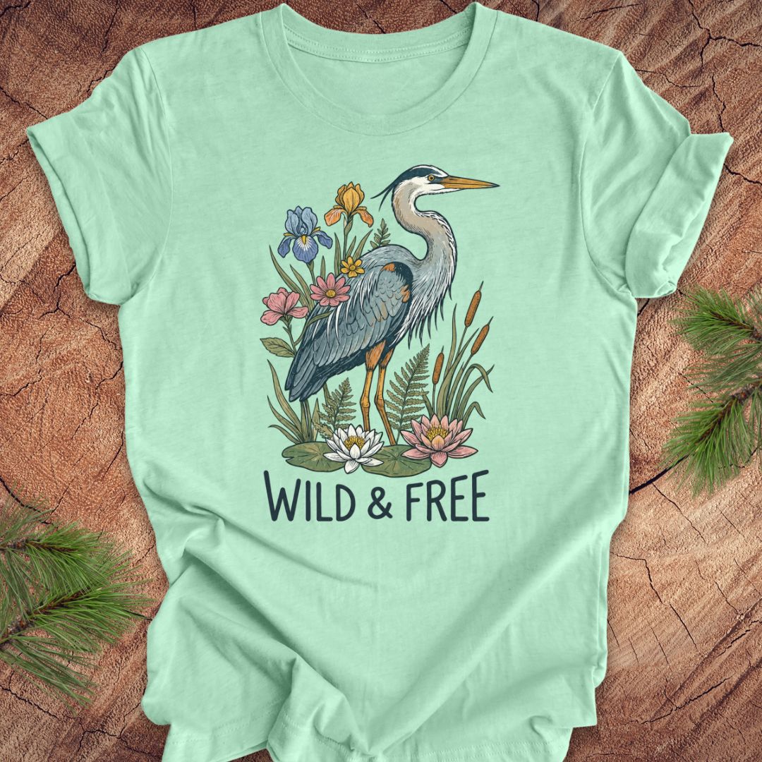 A mint green short-sleeve t-shirt with a graphic of a blue heron surrounded by colorful wildflowers and the text 'WILD & FREE' printed below it. The t-shirt is laid flat on a rustic wood background with pine needles in the corners.