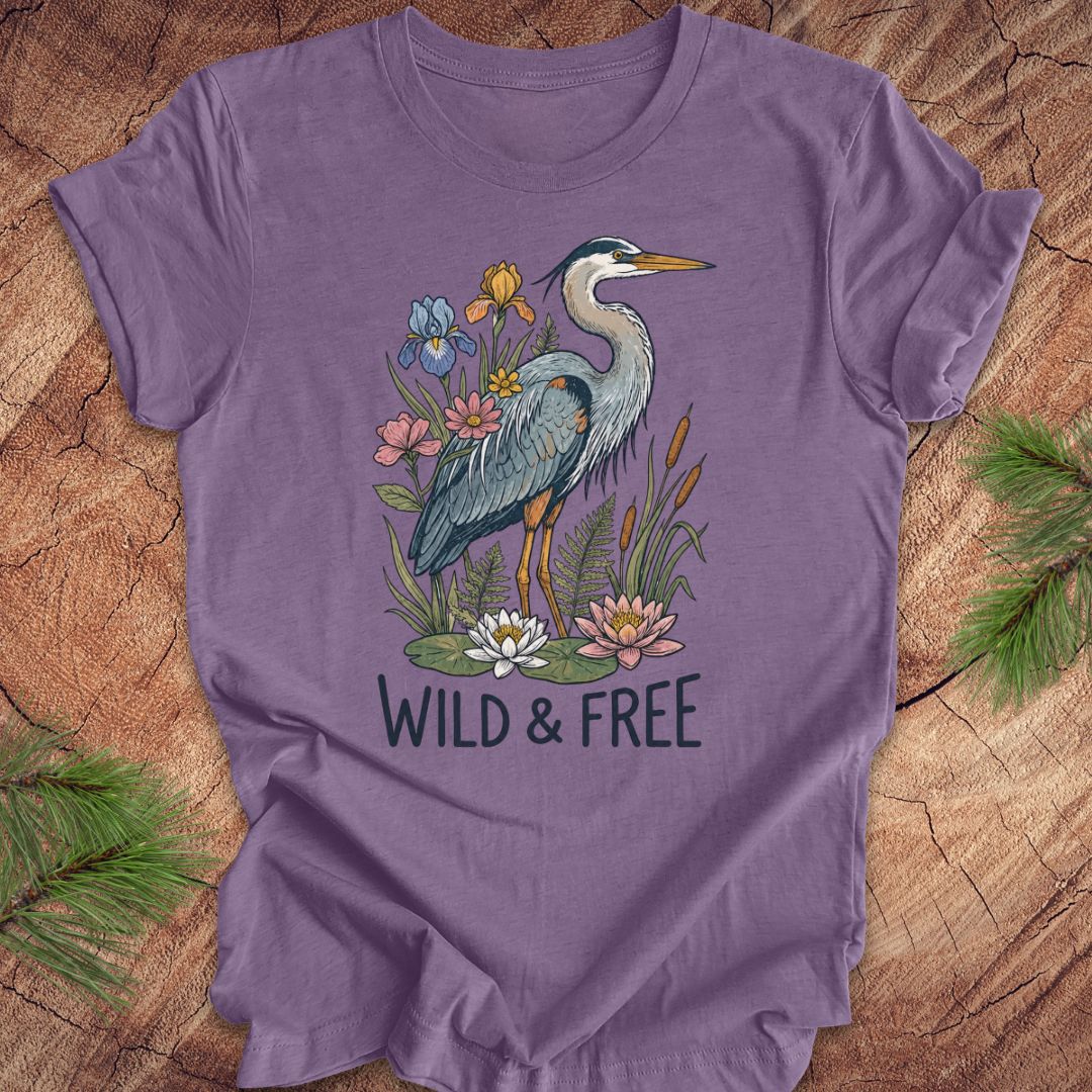 A purple short-sleeve t-shirt with a graphic of a blue heron surrounded by flowers and the text 'WILD & FREE', laid flat on a rustic wood background with pine branches.