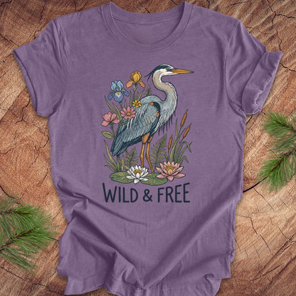 A purple short-sleeve t-shirt with a graphic of a blue heron surrounded by flowers and the text 'WILD & FREE', laid flat on a rustic wood background with pine branches.