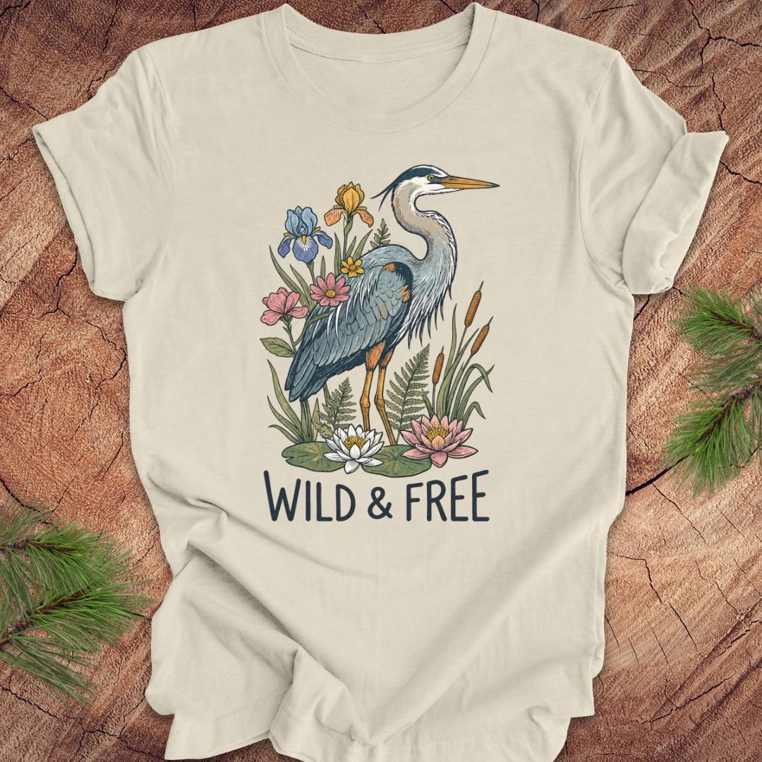 A cream-colored short-sleeve t-shirt with a graphic of a blue heron surrounded by wildflowers and the text 'WILD & FREE' printed below it. The t-shirt is laid flat on a rustic wood background with pine needles in the corners.