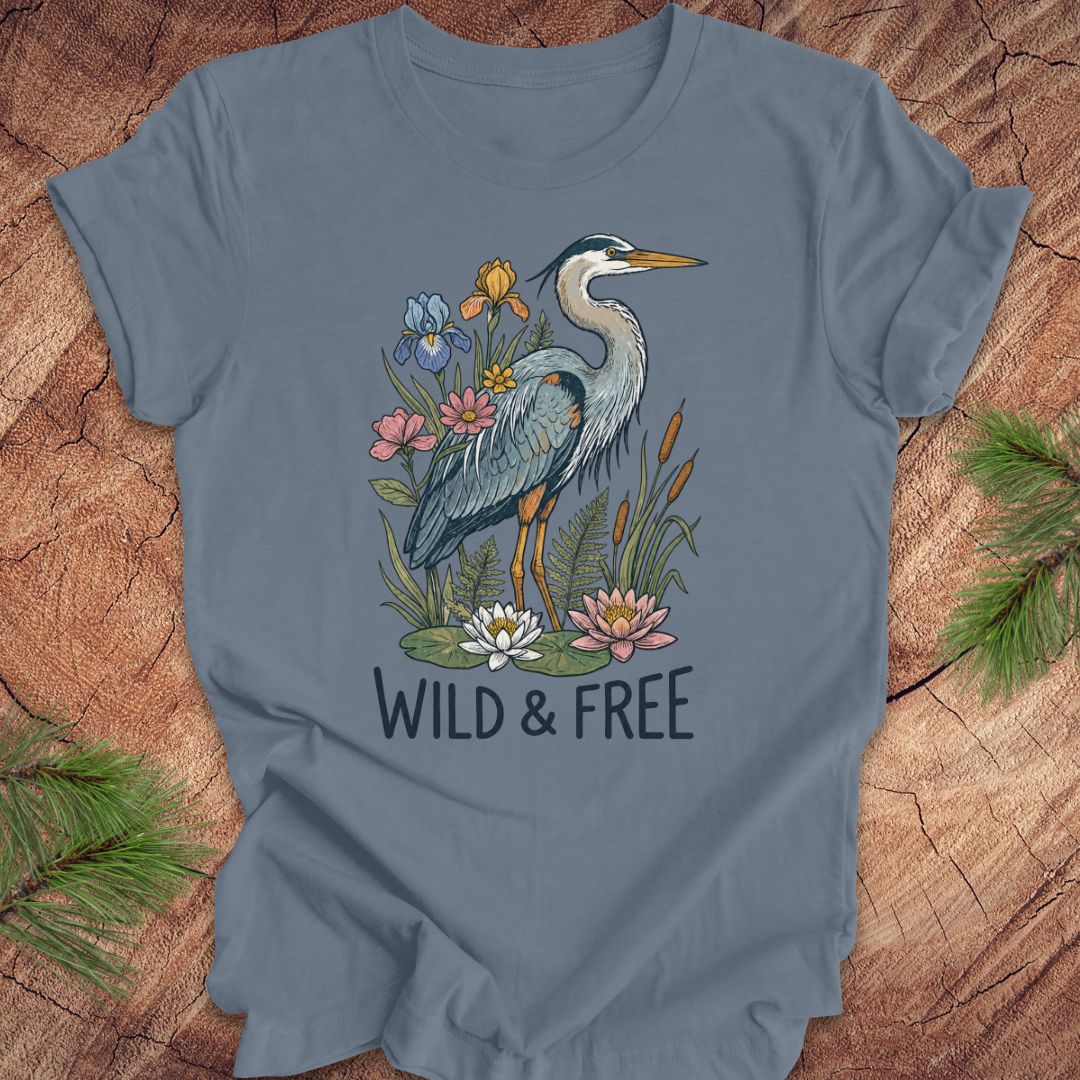 A slate blue short-sleeve t-shirt featuring a graphic of a blue heron surrounded by lilies and flowers, with the text 'WILD & FREE' printed below. The t-shirt is laid flat on a rustic wood background with pine branches in the corners.