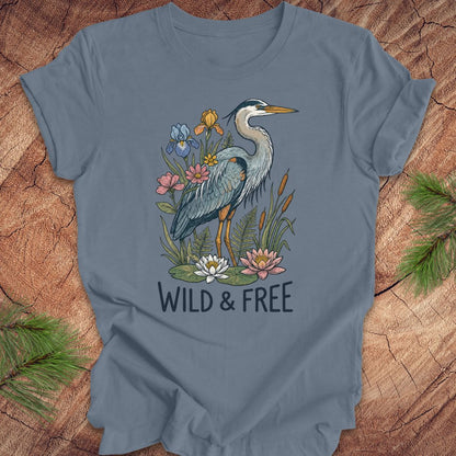 A slate blue short-sleeve t-shirt featuring a graphic of a blue heron surrounded by lilies and flowers, with the text 'WILD & FREE' printed below. The t-shirt is laid flat on a rustic wood background with pine branches in the corners.