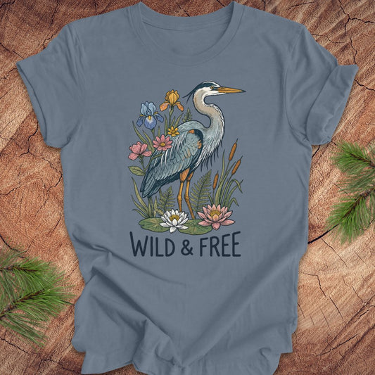 A slate blue short-sleeve t-shirt featuring a graphic of a blue heron surrounded by lilies and flowers, with the text 'WILD & FREE' printed below. The t-shirt is laid flat on a rustic wood background with pine branches in the corners.