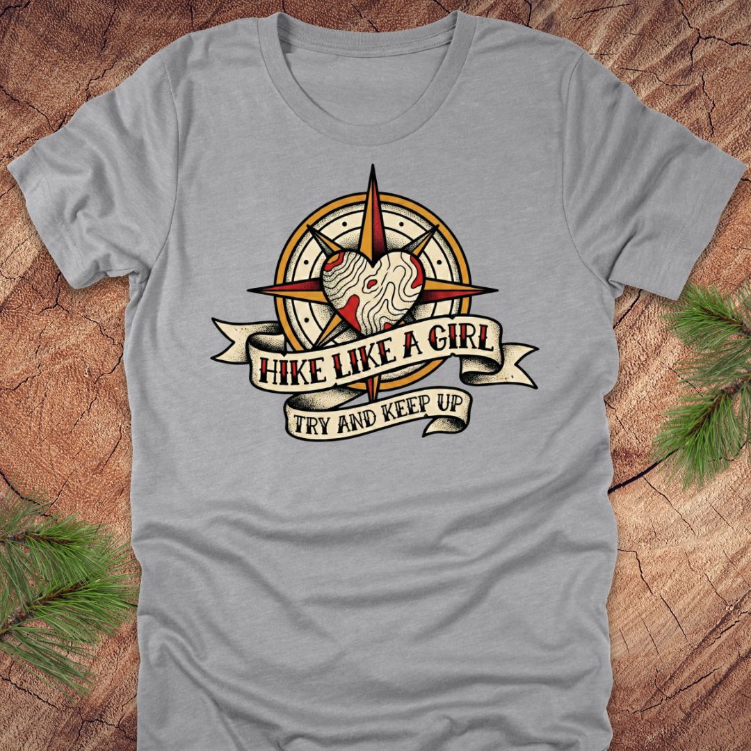 Gray t-shirt with a compass and heart design with text on a banner, 'hike like a girl. try and keep up' on a wood background.