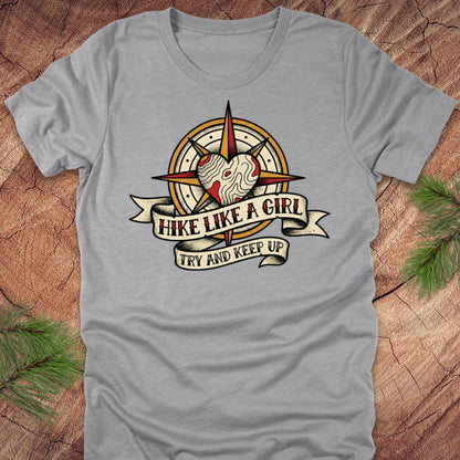 Gray t-shirt with a compass and heart design with text on a banner, 'hike like a girl. try and keep up' on a wood background.