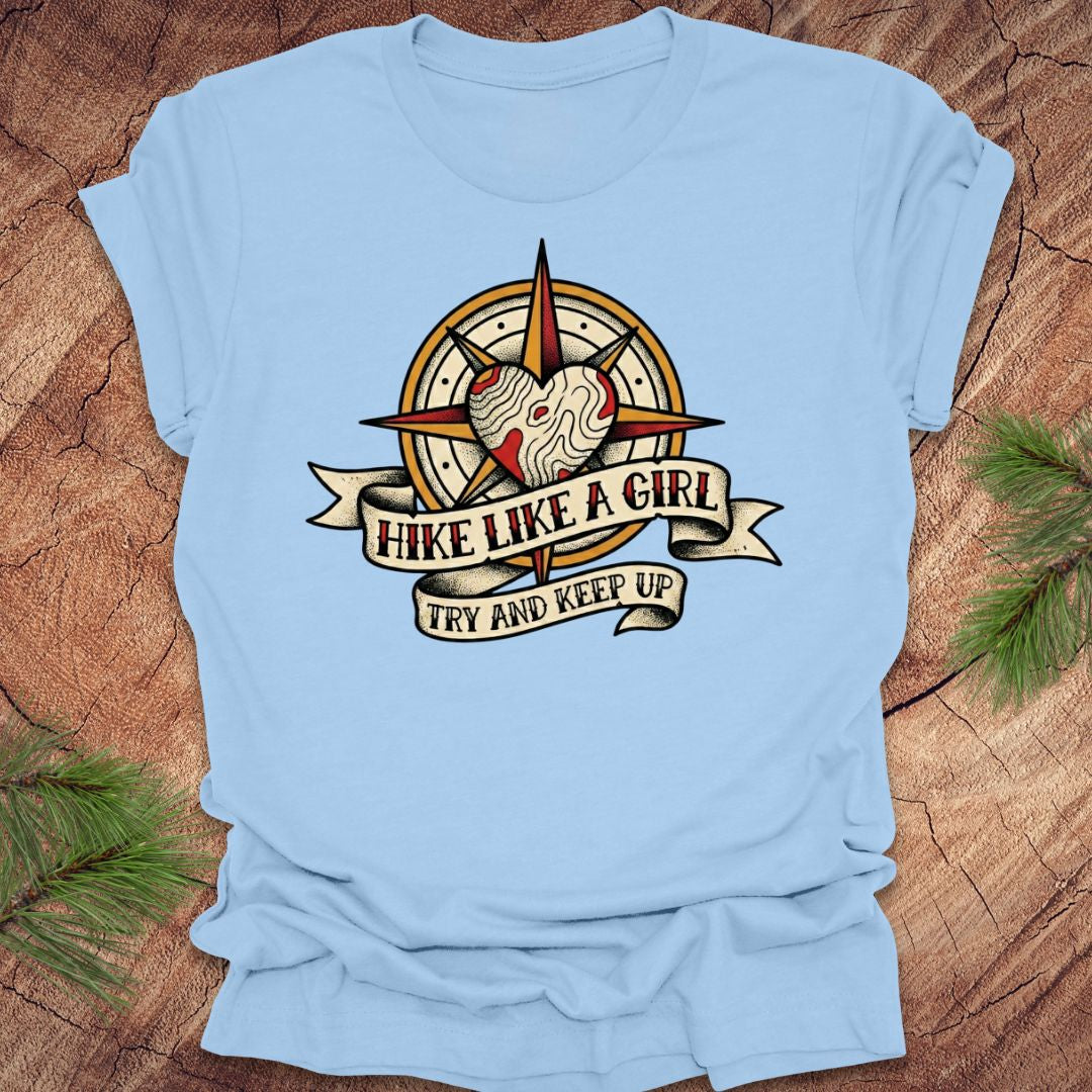 Baby blue t-shirt with a compass and heart design with text on a banner, 'hike like a girl. try and keep up' on a wood background.