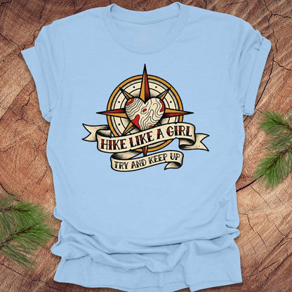 Baby blue t-shirt with a compass and heart design with text on a banner, 'hike like a girl. try and keep up' on a wood background.