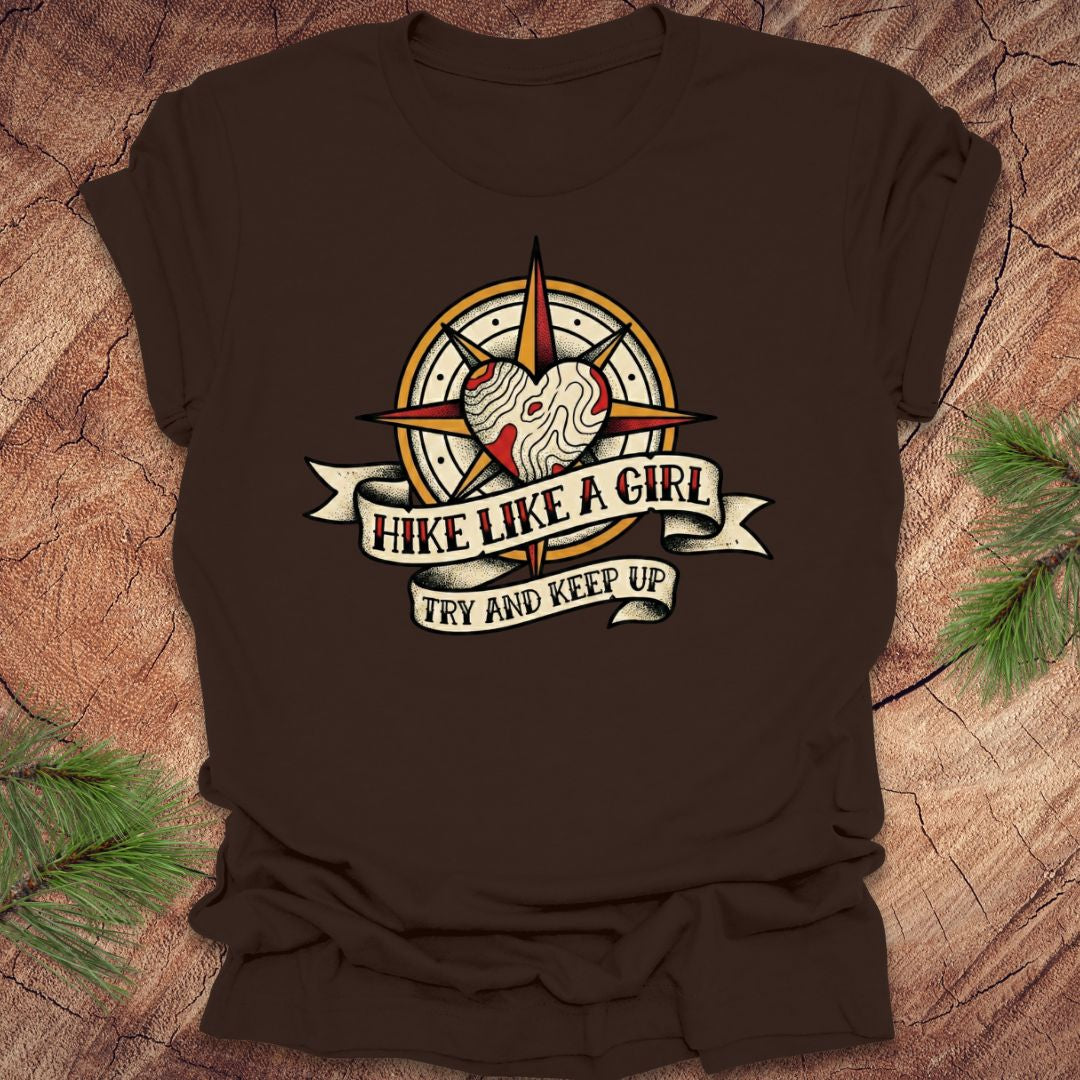 Brown t-shirt with a compass and heart design with text on a banner, 'hike like a girl. try and keep up' on a wood background.