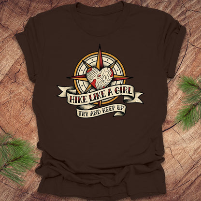 Brown t-shirt with a compass and heart design with text on a banner, 'hike like a girl. try and keep up' on a wood background.