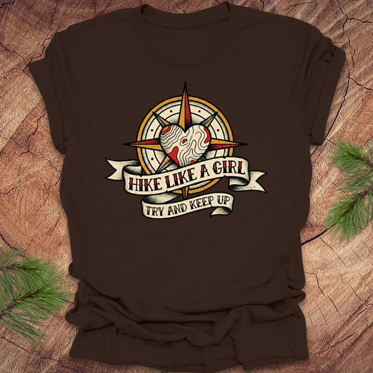 Brown t-shirt with a compass and heart design with text on a banner, 'hike like a girl. try and keep up' on a wood background.