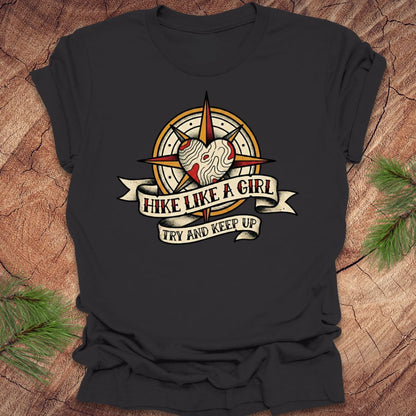 Dark gray t-shirt with a compass and heart design with text on a banner, 'hike like a girl. try and keep up' on a wood background.