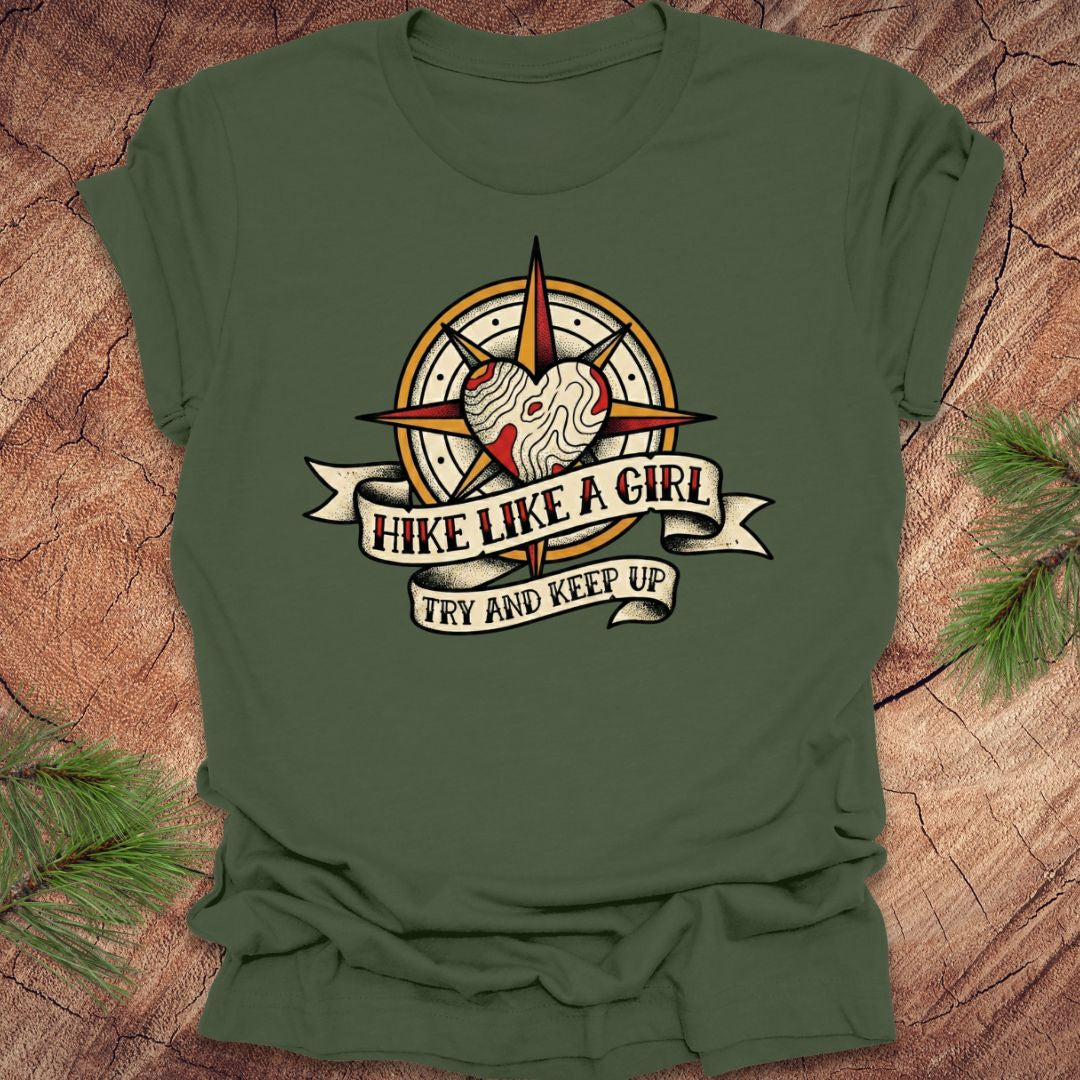 Green t-shirt with a compass and heart design with text on a banner, 'hike like a girl. try and keep up' on a wood background.