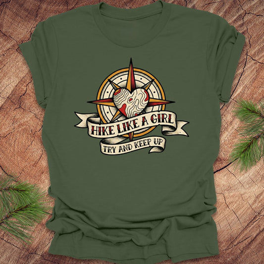 Green t-shirt with a compass and heart design with text on a banner, 'hike like a girl. try and keep up' on a wood background.