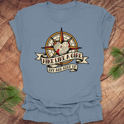 Steel blue t-shirt with a compass and heart design with text on a banner, 'hike like a girl. try and keep up' on a wood background.