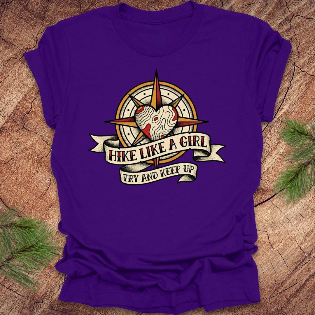 Purple t-shirt with a compass and heart design with text on a banner, 'hike like a girl. try and keep up' on a wood background.