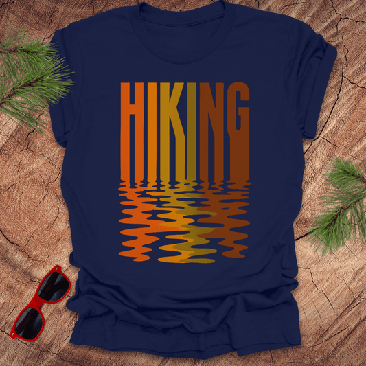 Navy blue t-shirt with the text "Hiking" graphic reflecting in earth toned colored water, on a wood background.