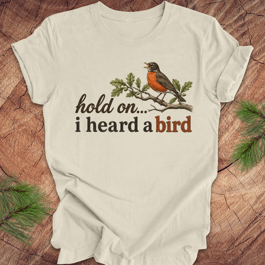 Beige t-shirt with a robin graphic and text 'hold on... i heard a bird' on a wooden surface.