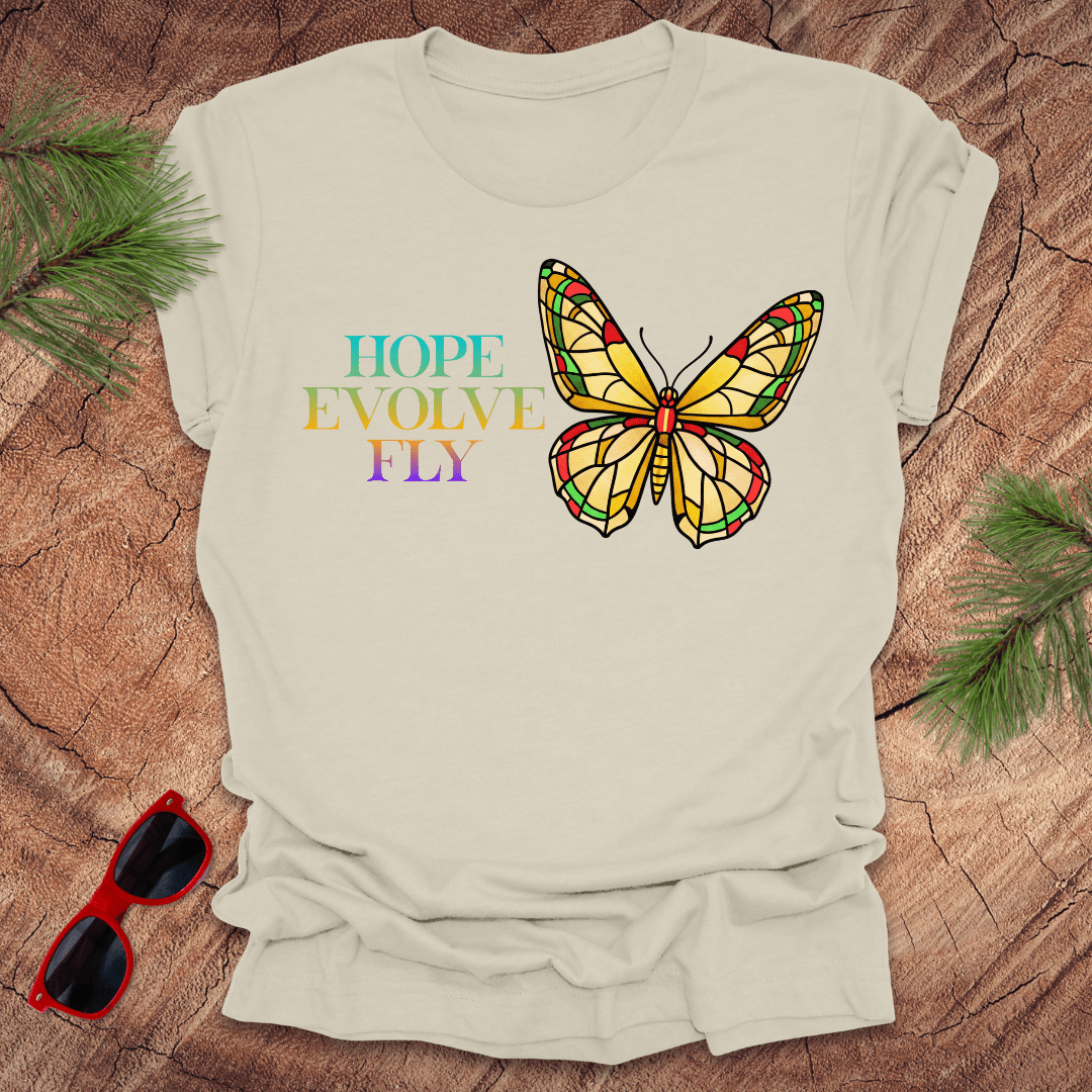 Beige t-shirt with a colorful butterfly graphic and text on a wooden surface with sunglasses and greenery.
