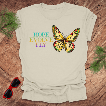 Beige t-shirt with a colorful butterfly graphic and text on a wooden surface with sunglasses and greenery.
