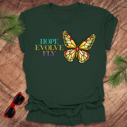 Green t-shirt with a colorful butterfly graphic and text on a wooden surface with sunglasses and pine branches.