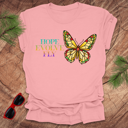 Pink t-shirt with a butterfly graphic and text on a wooden surface with sunglasses and greenery.