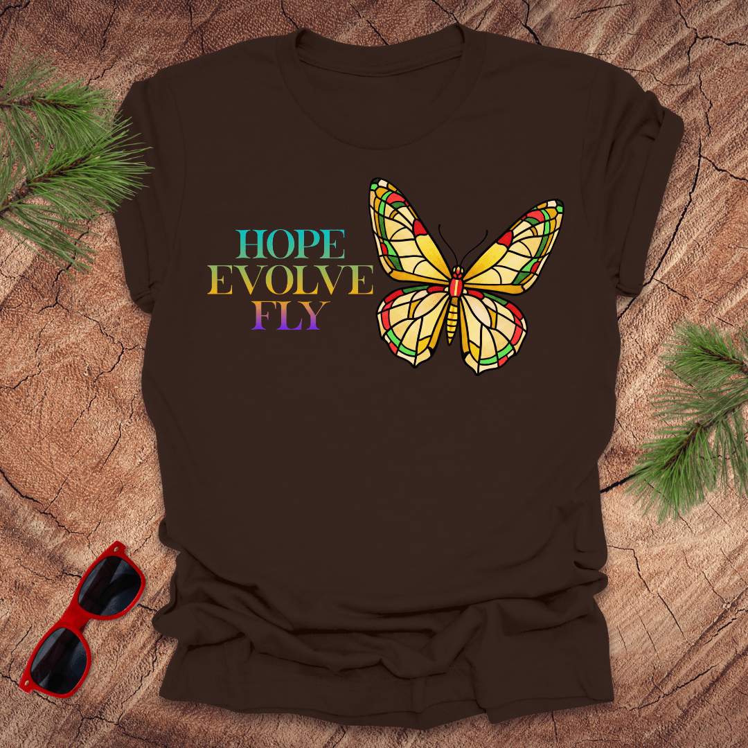 Brown t-shirt with a colorful butterfly graphic and text on a wooden surface with sunglasses and greenery.