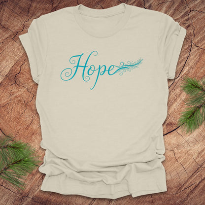 Beige t-shirt with hand drawn text, 'Hope' in turquoise on a wooden surface with greenery.