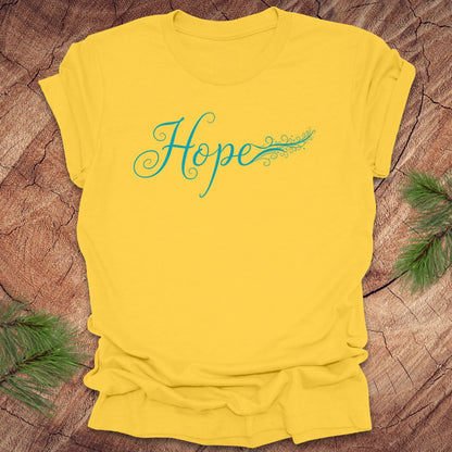 Yellow t-shirt with hand drawn text, 'Hope' in turquoise on a wooden surface with greenery.