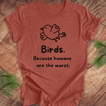 Clay colored t-shirt with a bird and text, 'birds. because humans are the worst.' on a wood background. 