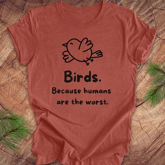 Clay colored t-shirt with a bird and text, 'birds. because humans are the worst.' on a wood background. 