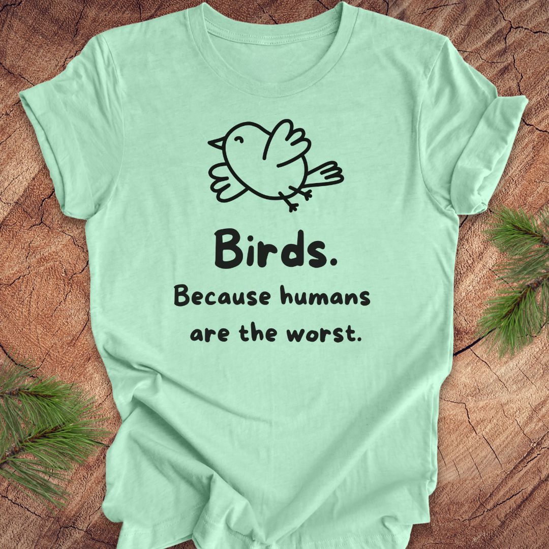 Mint green t-shirt with a bird and text, 'birds. because humans are the worst.' on a wood background. 