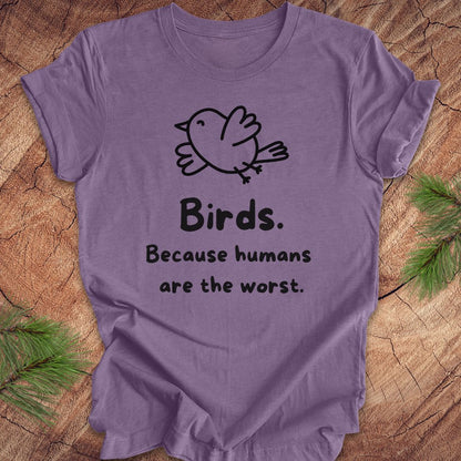 Purple t-shirt with a bird and text, 'birds. because humans are the worst.' on a wood background. 