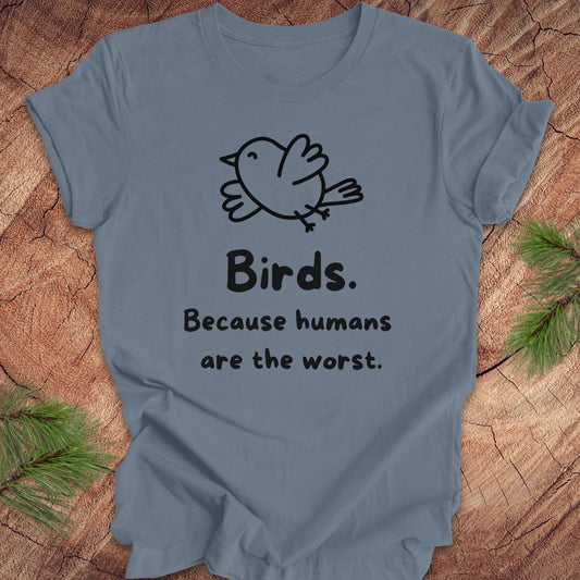 Blue t-shirt with a bird and text, 'birds. because humans are the worst.' on a wood background. 