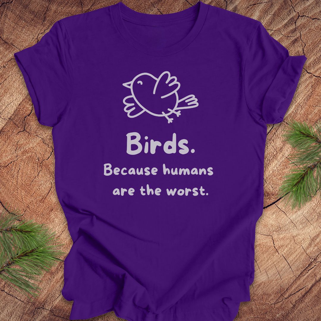 Purple t-shirt with a bird and text, 'birds. because humans are the worst.' on a wood background. 